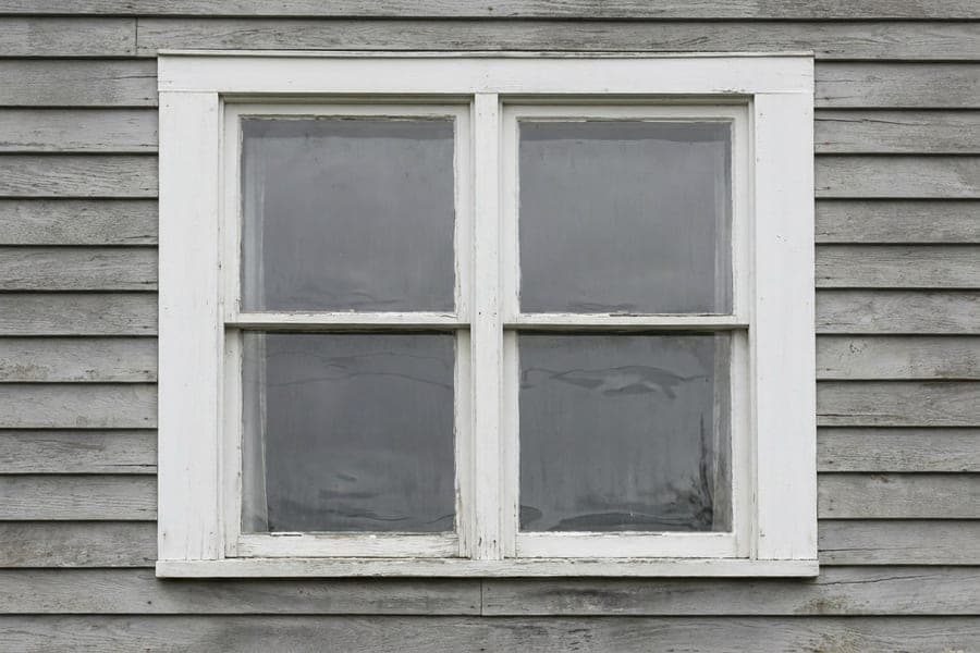 A white double hung window with flat casing on grey clapboard siding.