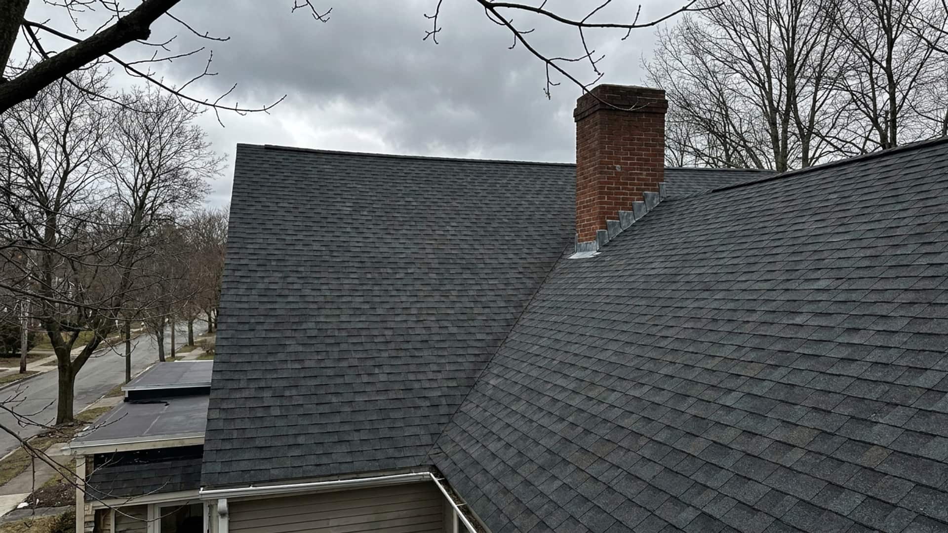A steep asphalt shingle roof with a brick chimney and step flashing on an older colonial house.